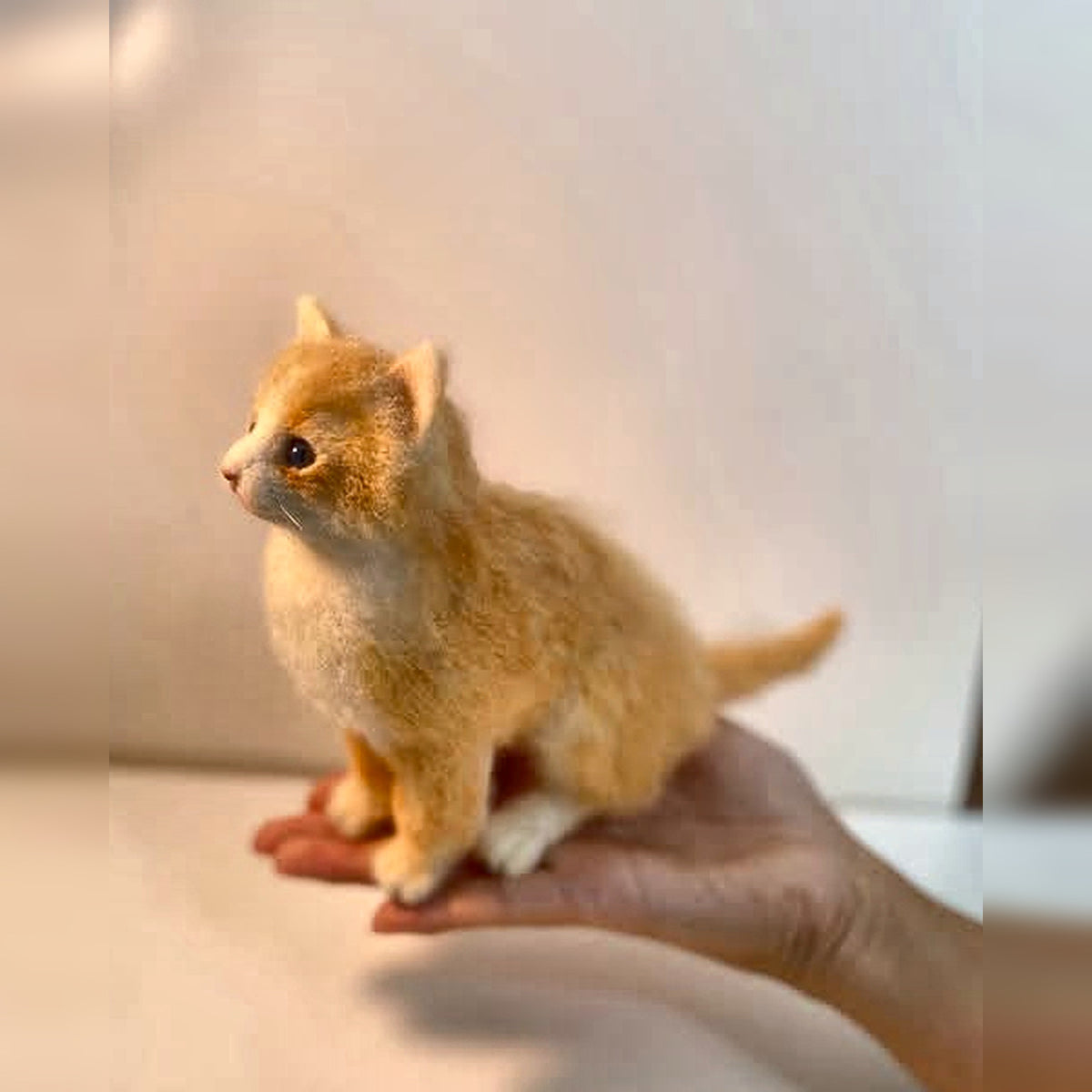 Hand holding a small finished ginger felt kitten sculpture to show its scale; square image