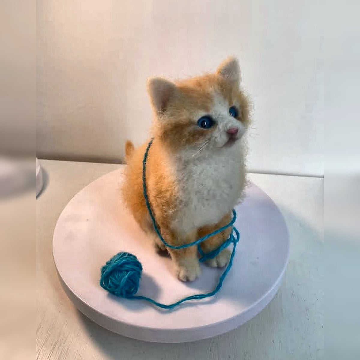 Three-quarter view of the ginger felt kitten showing its round eyes, pink nose and white chest; square image